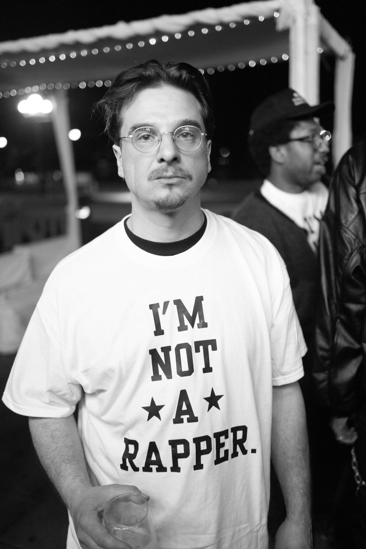 A man wearing round glasses and a white graphic t-shirt with the text 'I'M NOT A RAPPER.' in black, standing outdoors at night with string lights in the background.