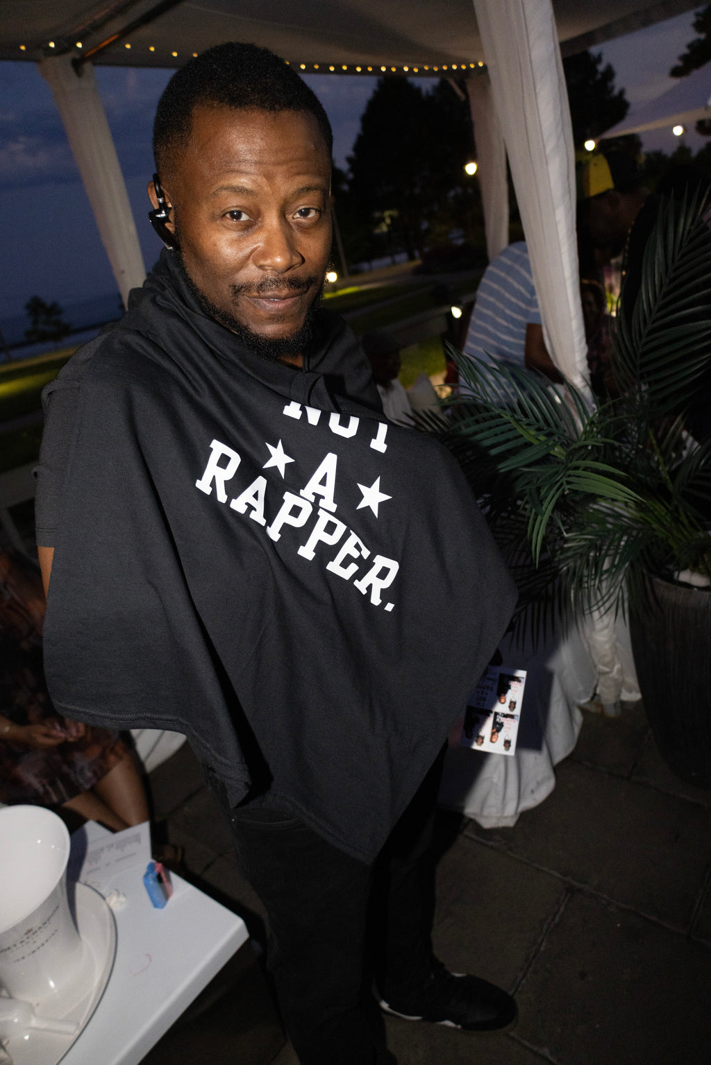 A man wearing a black hoodie with 'NOT A RAPPER.' printed in white across the chest, standing at an outdoor evening event under a tent.